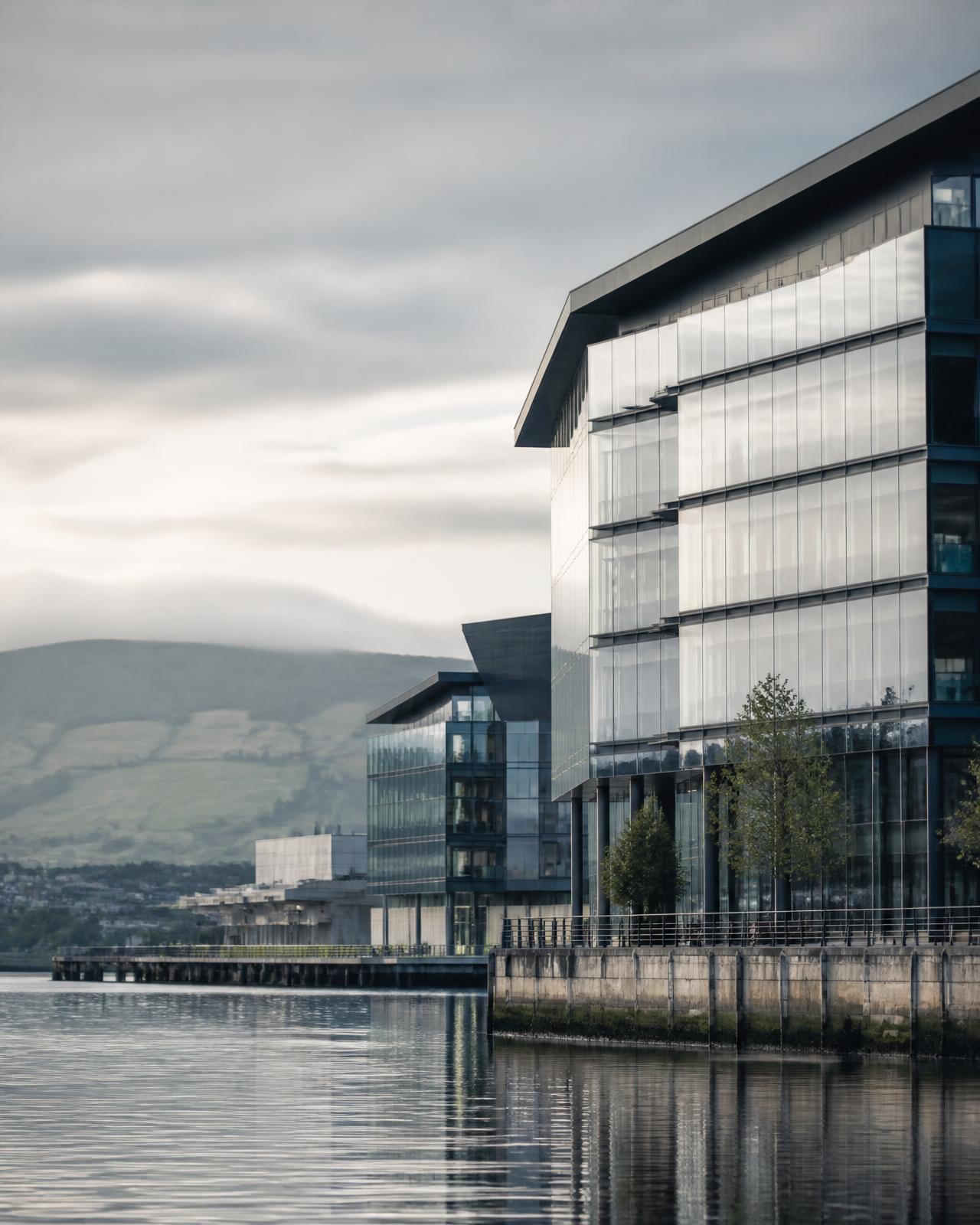 Modern glass office buildings on an Irish waterfront with green hills behind in soft morning light